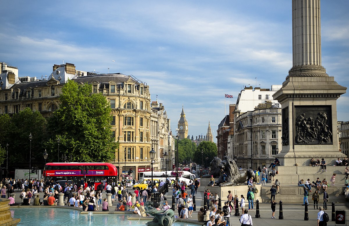 Trafalgar square in London on a summer's day. Dozens of people are sitting around the fountain and on a large obelisk monument.