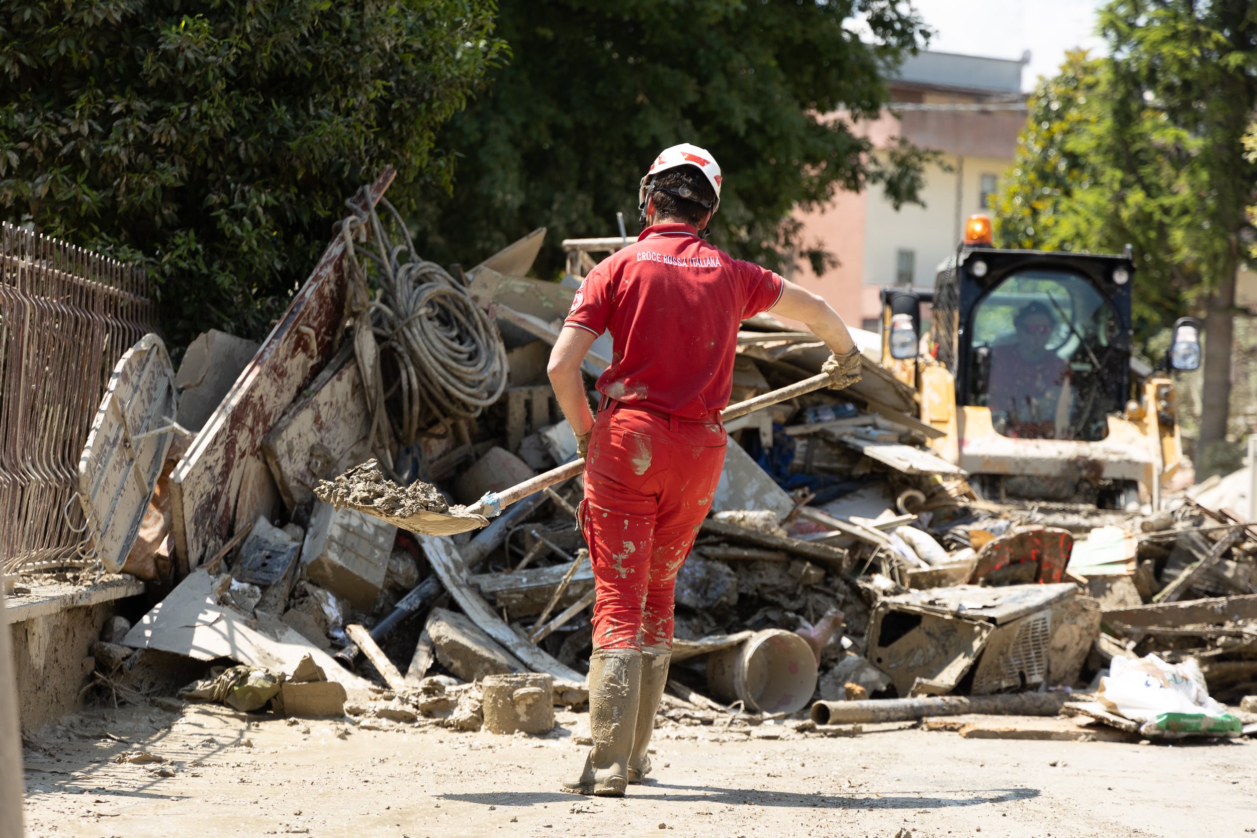 A red cross worker wearing red pants, a red shirt, a helmet and gumboots walks on street with a shovel towards a large pile of flood debris.
