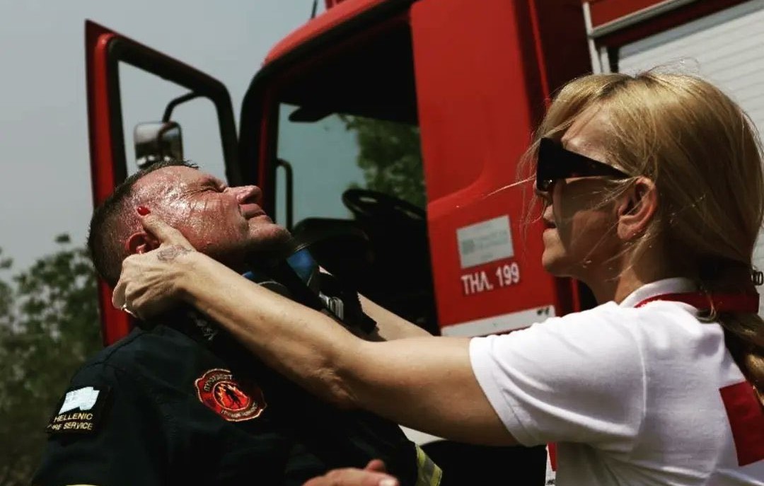 A Greek firefighter receives care from a Red Cross worker. The firefighter is sweating and grimacing and the Red Cross worker places her hands on his face.