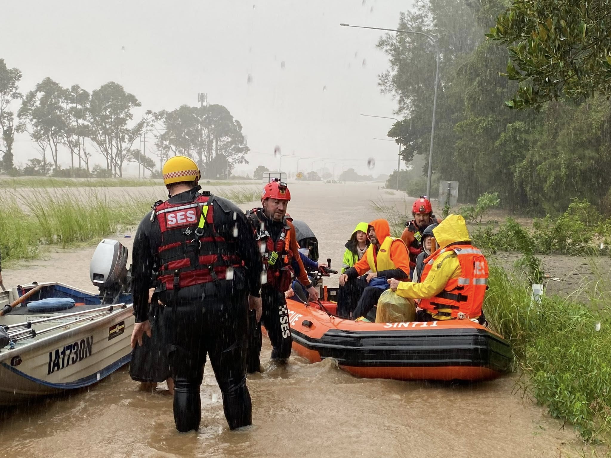 Two men in wetsuits are in knee deep brown water pushing two inflatable boats with people in them.