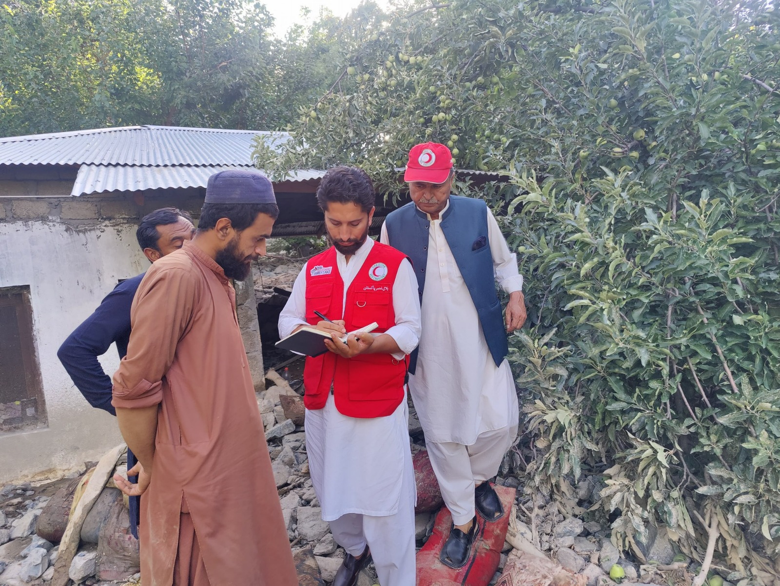 A man from the Pakistan Red Cross wearing a red bib speaks to a Pakistani man in front of a damaged house.