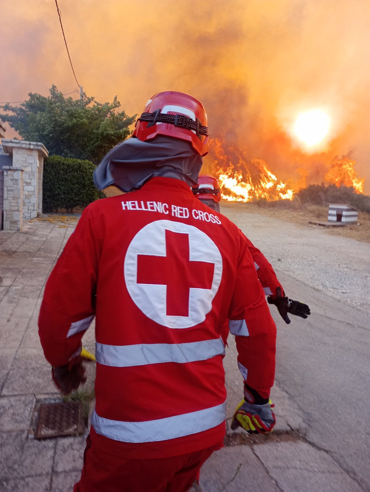 A person in a red cross red jump suit with 'Hellenic Red Cross' on the back and a large white cross walks toward a large wildfire up the road