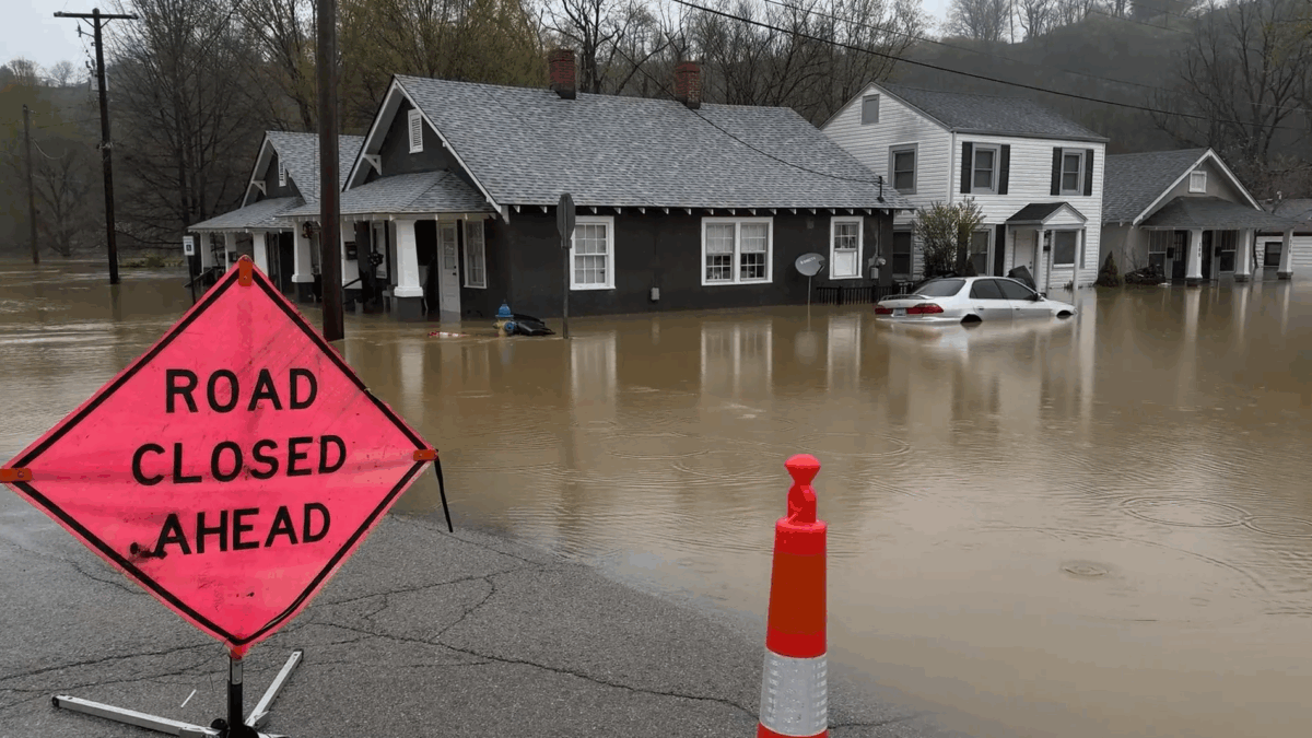 An image of a flooded road with a sign that says 'road closed ahead’, a house with brown water up to its windows and a submerged car.