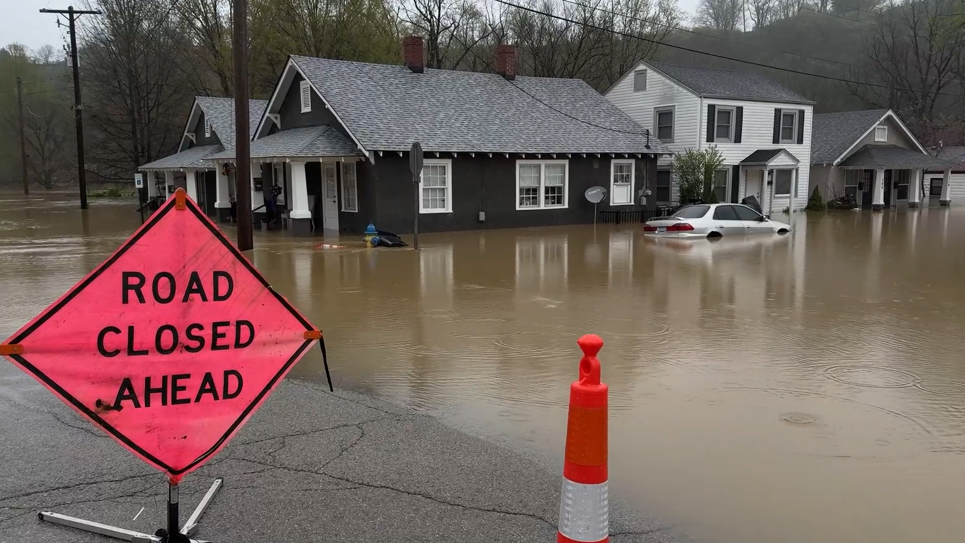 An image of a flooded road with a sign that says 'road closed ahead’, a house with brown water up to its windows and a submerged car.