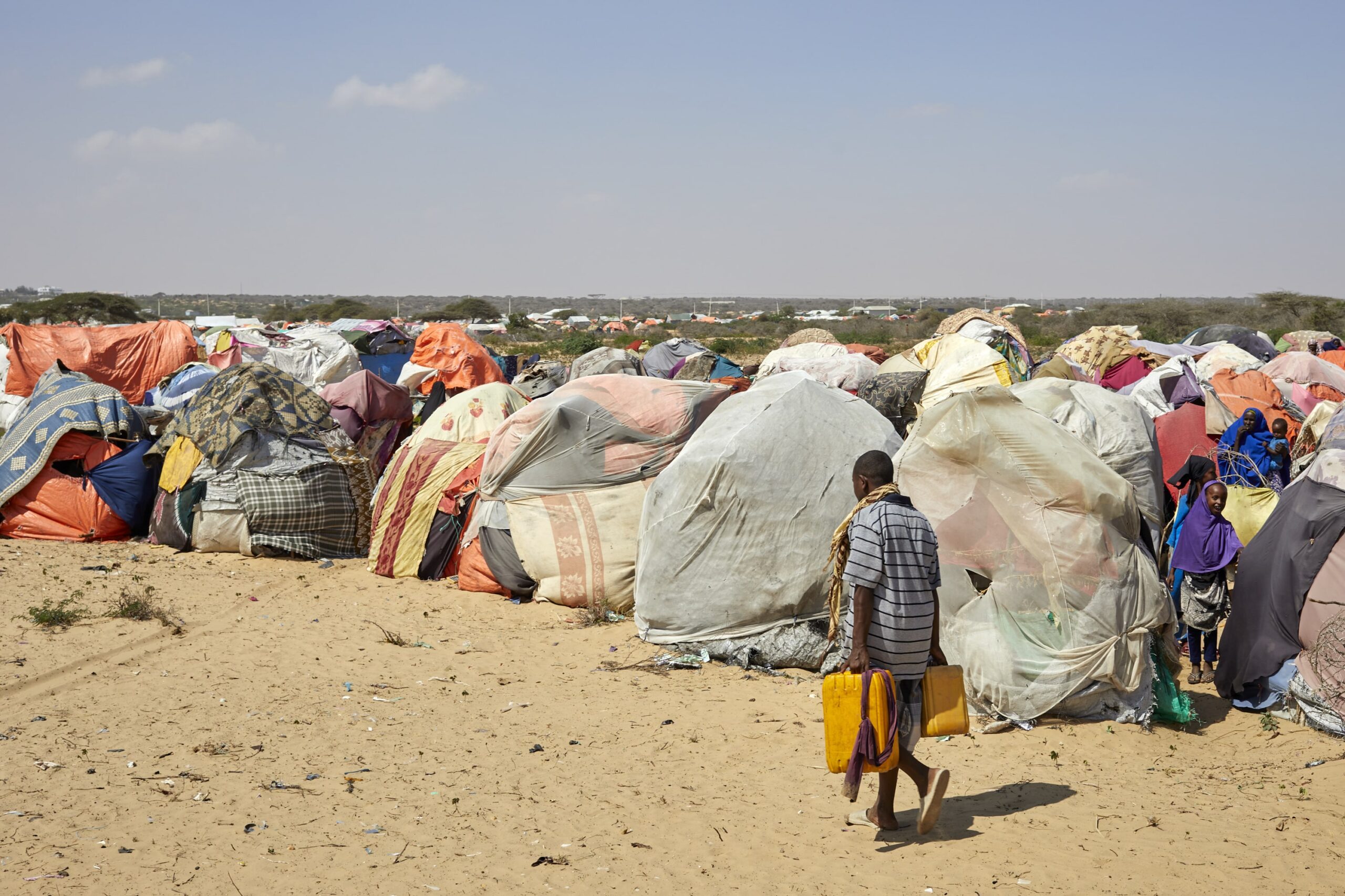 A refugee carrying two yellow water containers walks in front of a refugee camp in Somalia with tents surrounded by sand and shrubs.