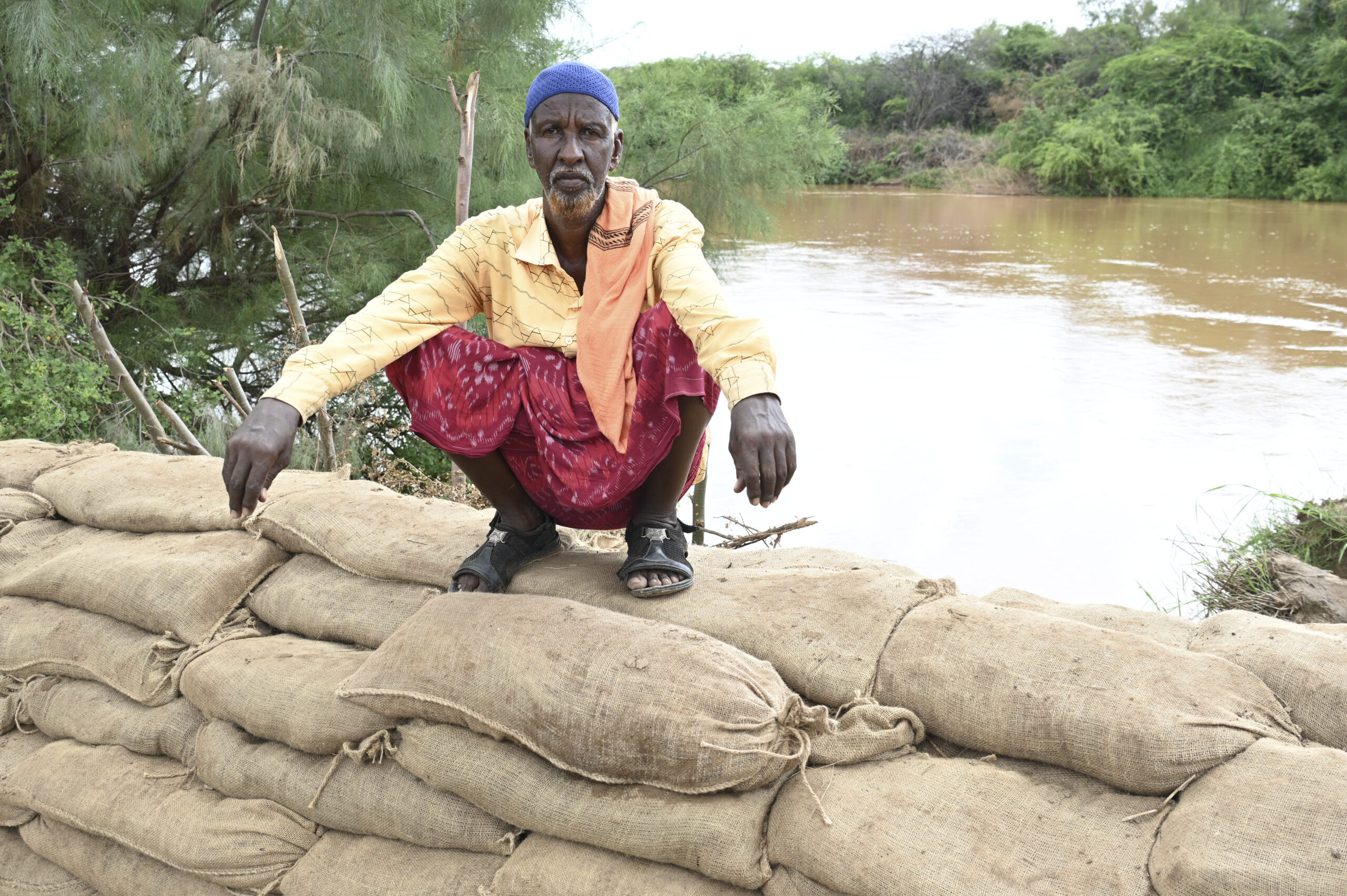 A man sits on top of a pile of sandbags in Somalia with a river behind. Very heavy rainfall is on the way.