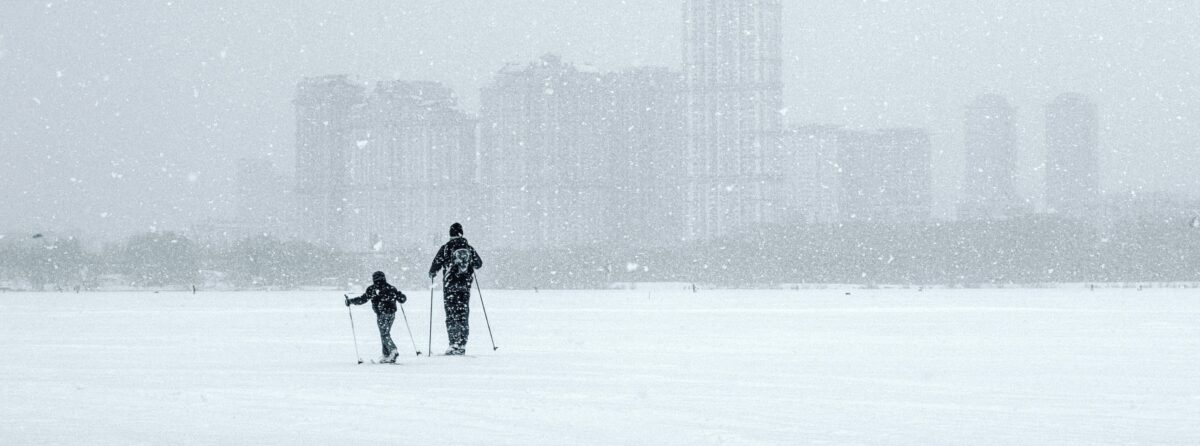 Two people cross country skiing in the snow in front of a city with grey skies above