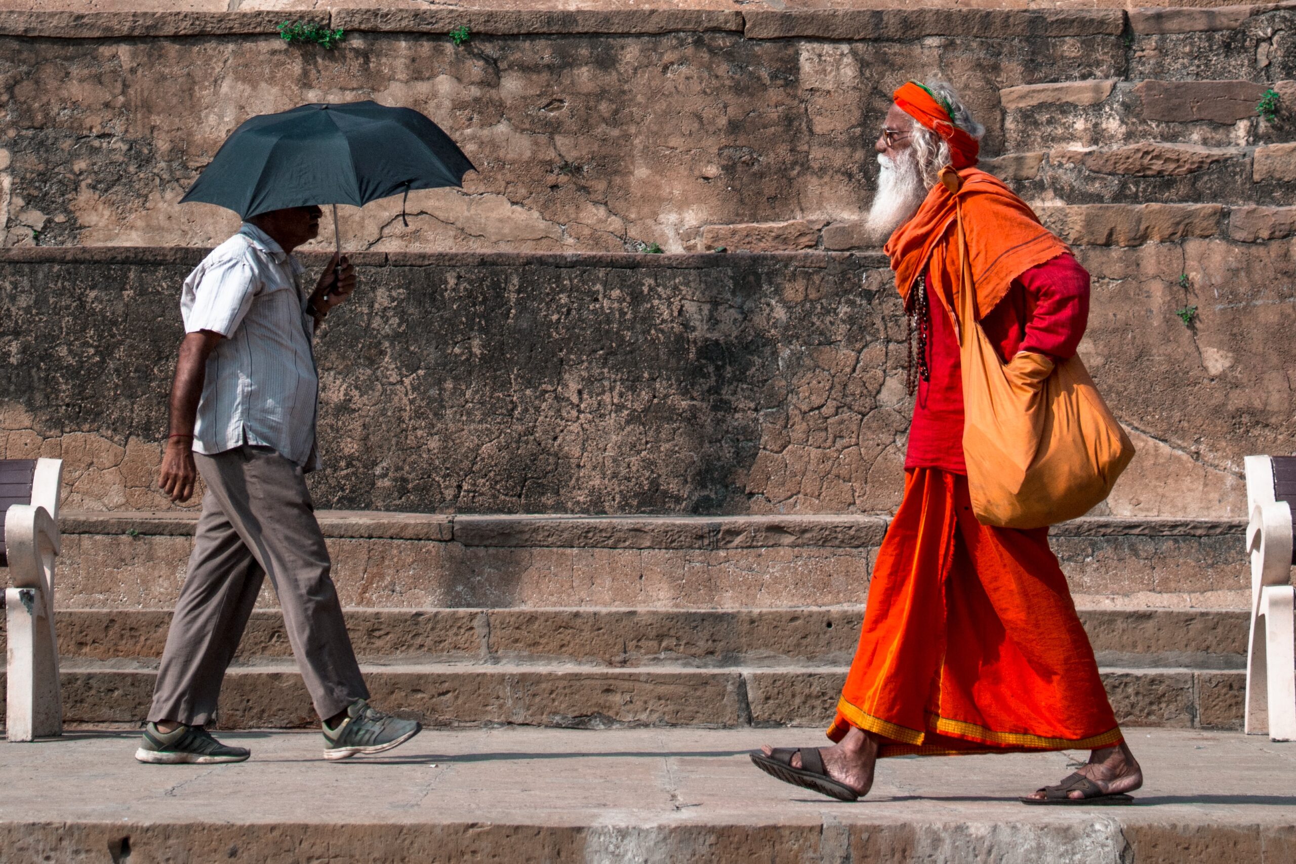 Two men walk past each other in India. One man wears a pants and shirts and holds an umbrella and the other man wears orange robes.