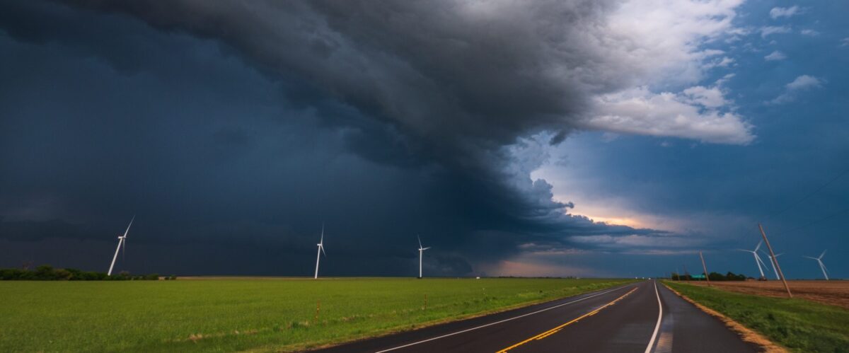 A dark storm travels across the sky over wind turbines, a road and grassy fields.